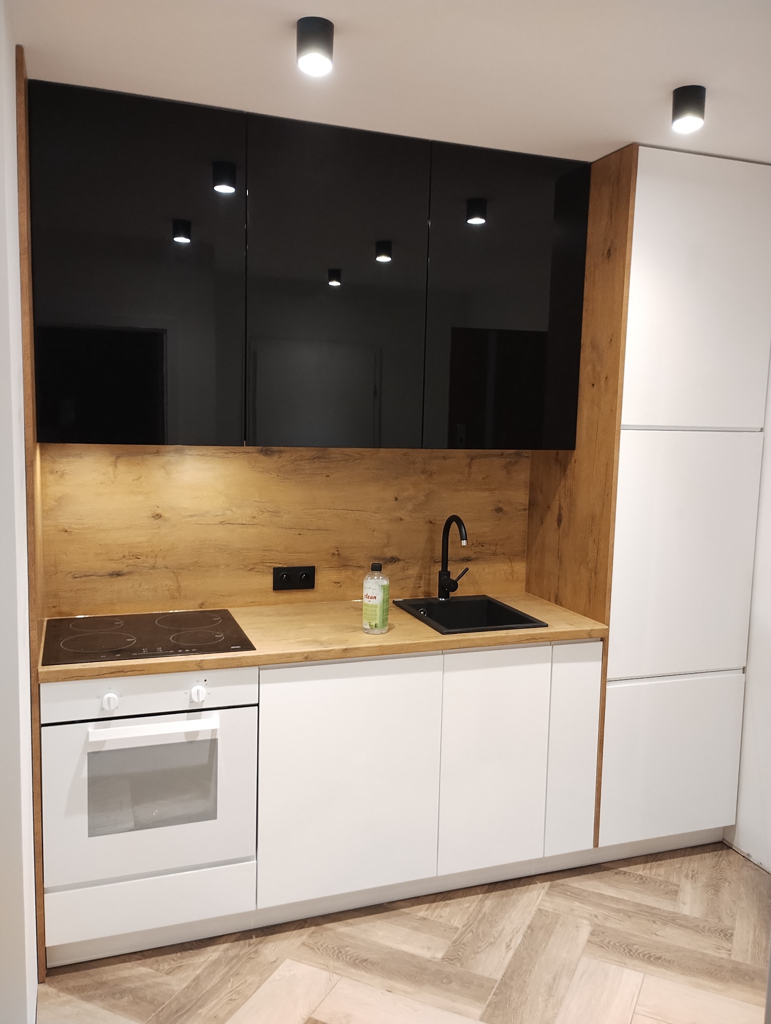 Nowoczesna kuchnia z białymi szafkami dolnymi, czarnymi szafkami górnymi na wysoki połysk, blatem imitującym drewno, czarnym zlewem i baterią oraz płytą indukcyjną i piekarnikiem w zabudowie.