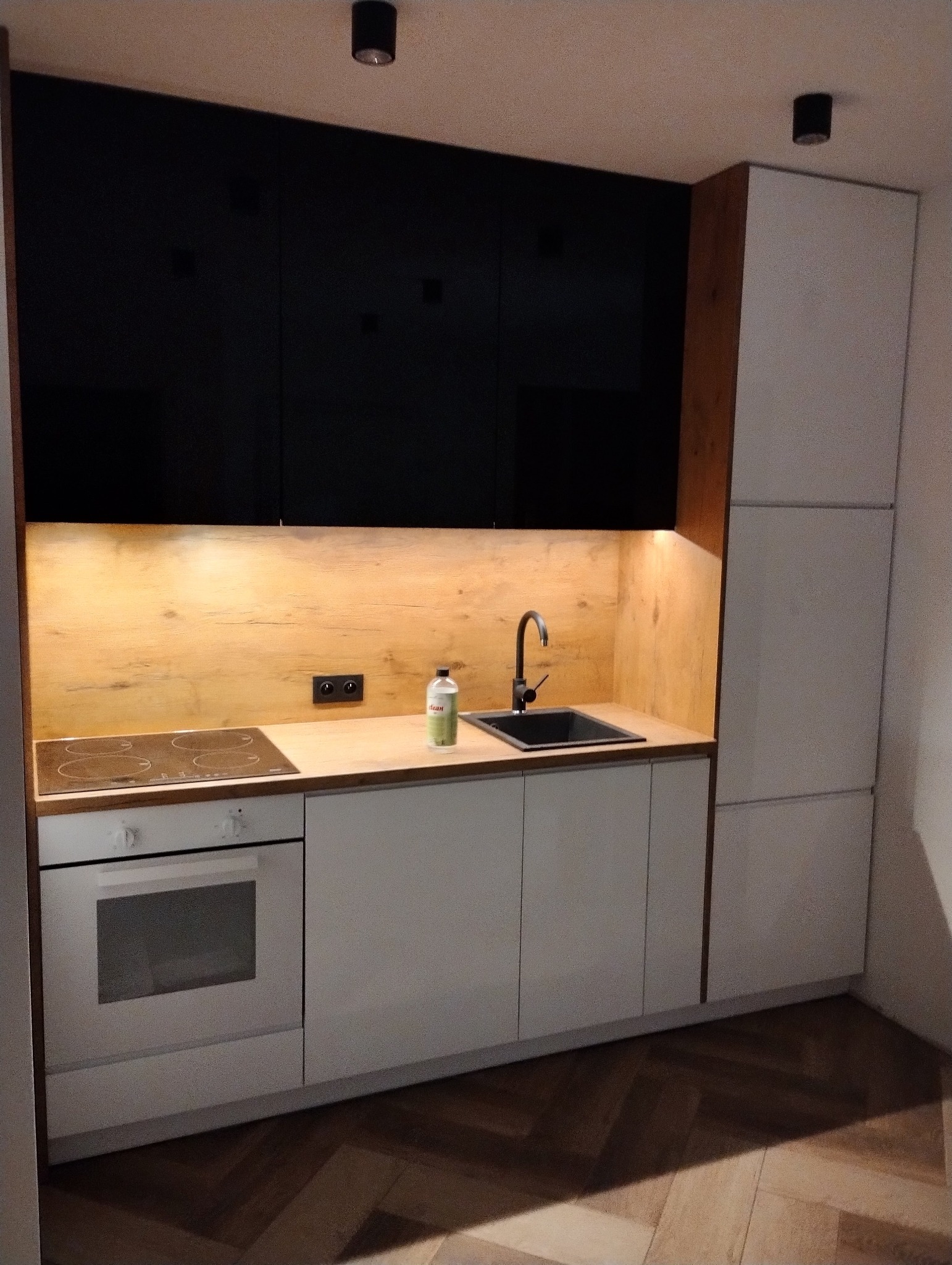 Nowoczesna kuchnia z czarnymi szafkami górnymi i jasnymi frontami dolnymi, drewniany panel nad blatem, indukcyjna płyta grzewcza i czarny zlew z baterią.