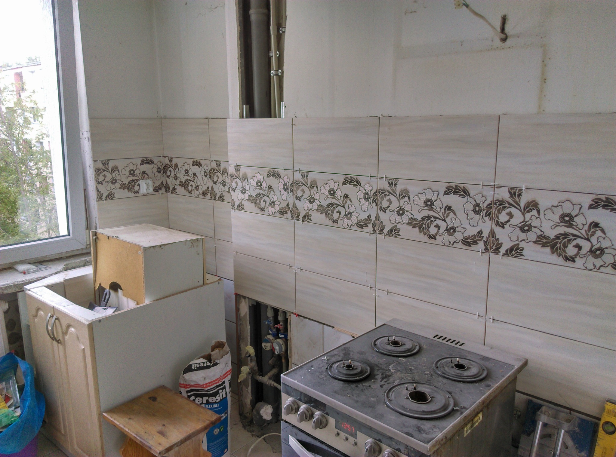 Remont kuchni w trakcie: częściowo położone płytki z motywem kwiatowym na ścianie, odsłonięte rury, stara kuchenka gazowa i worek z klejem do płytek.