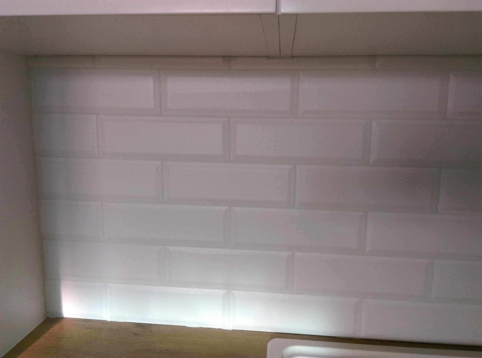 Białe, błyszczące płytki typu metro ułożone na ścianie w kuchni, widoczne fugi i oświetlenie LED pod szafkami, blat drewnopodobny.