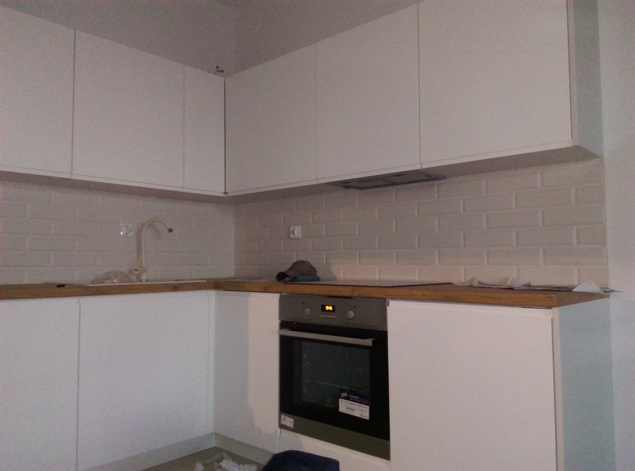 Nowoczesna kuchnia z białymi szafkami, drewnianym blatem i białymi płytkami imitującymi cegłę, widok na piekarnik i fragment zlewu.