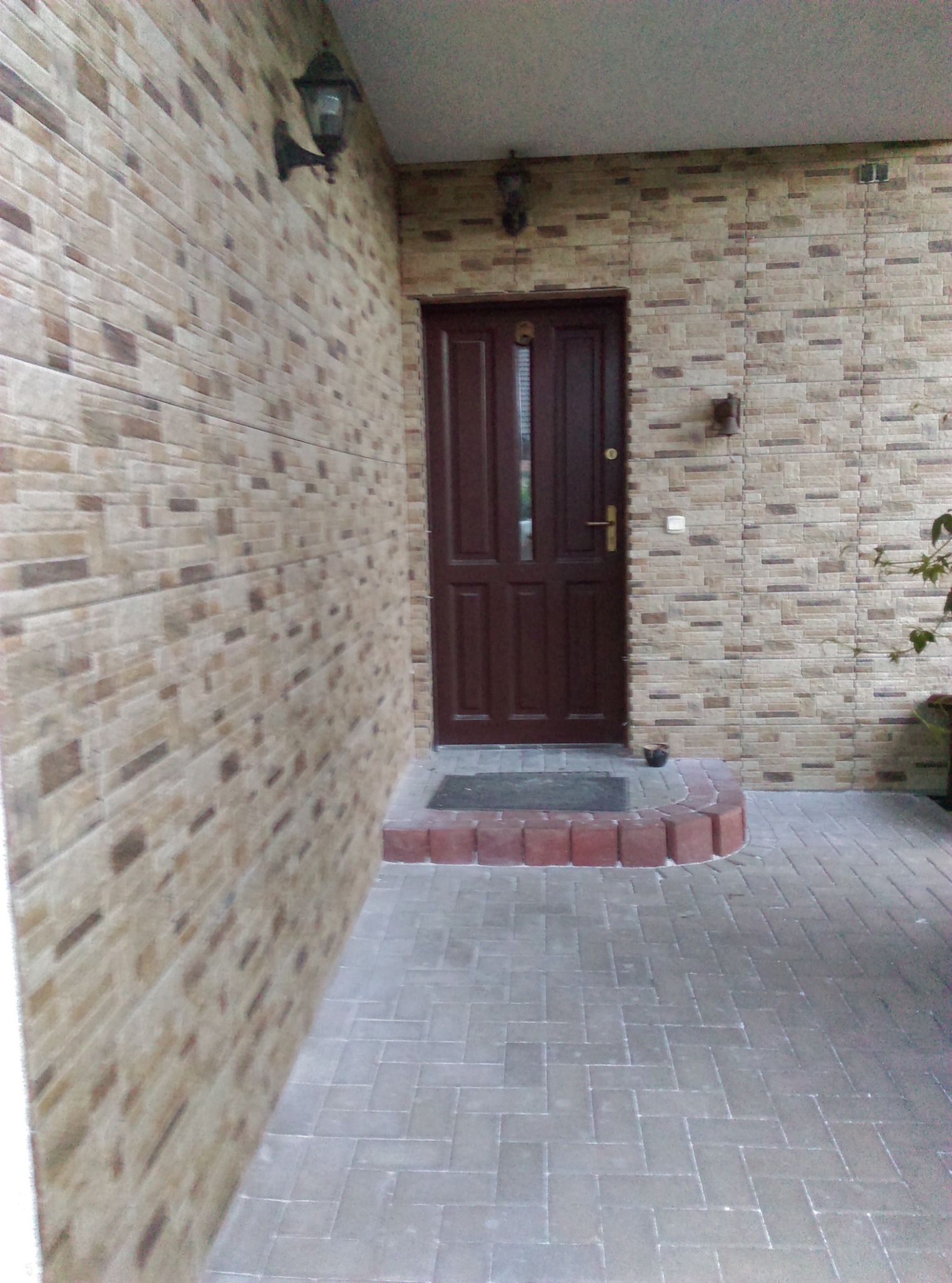 Zewnętrzna ściana budynku wykończona beżową, imitującą cegłę okładziną, brązowe drzwi wejściowe z dwoma przeszkleniami, oświetlenie zewnętrzne w stylu retro, chodnik wyłożony szarą kostką brukową.