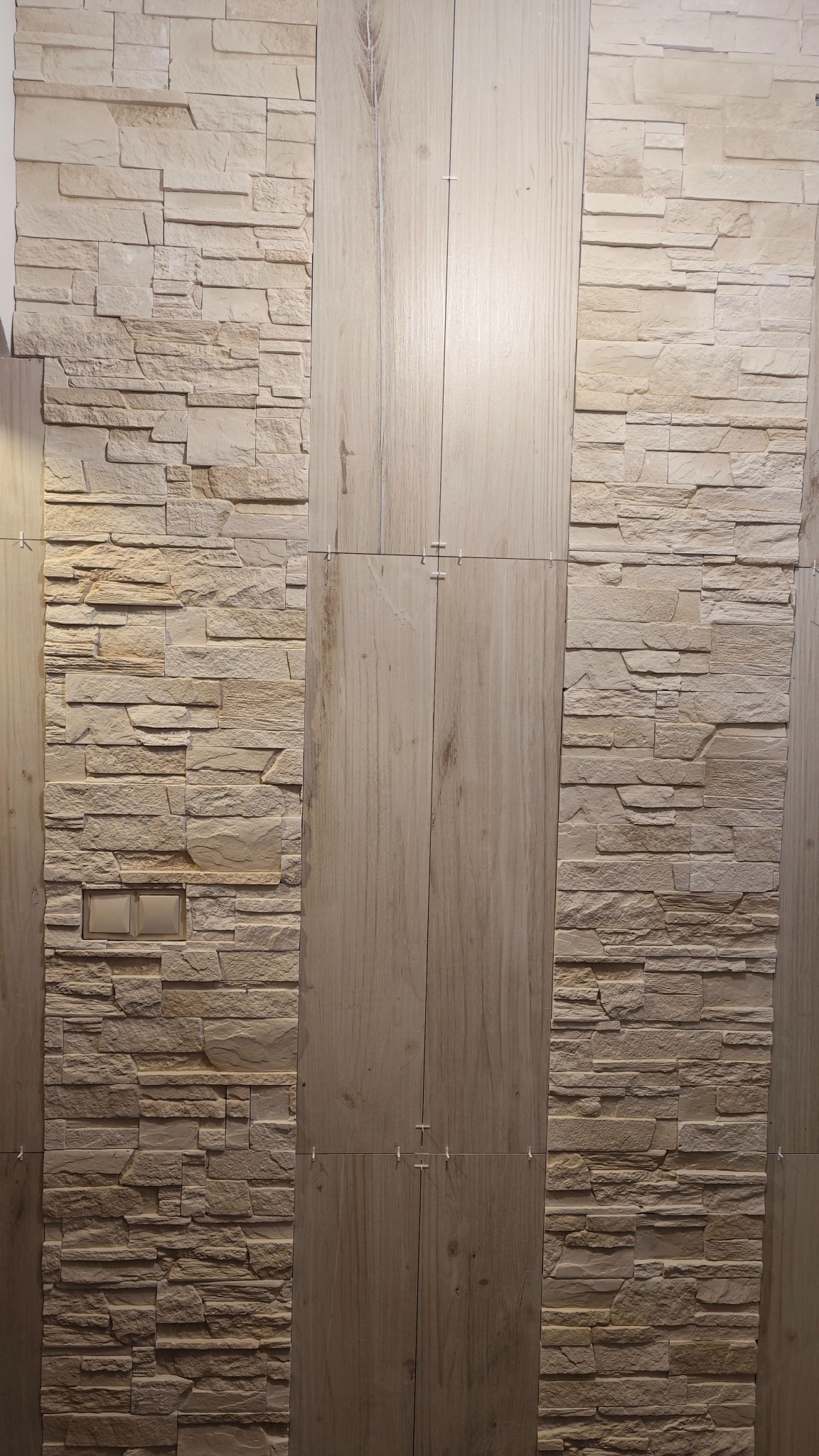 Ściana wykończona kombinacją płytek imitujących kamień i drewno, z widocznymi krzyżykami dystansowymi, sugerującymi świeżo zakończony montaż płytek.