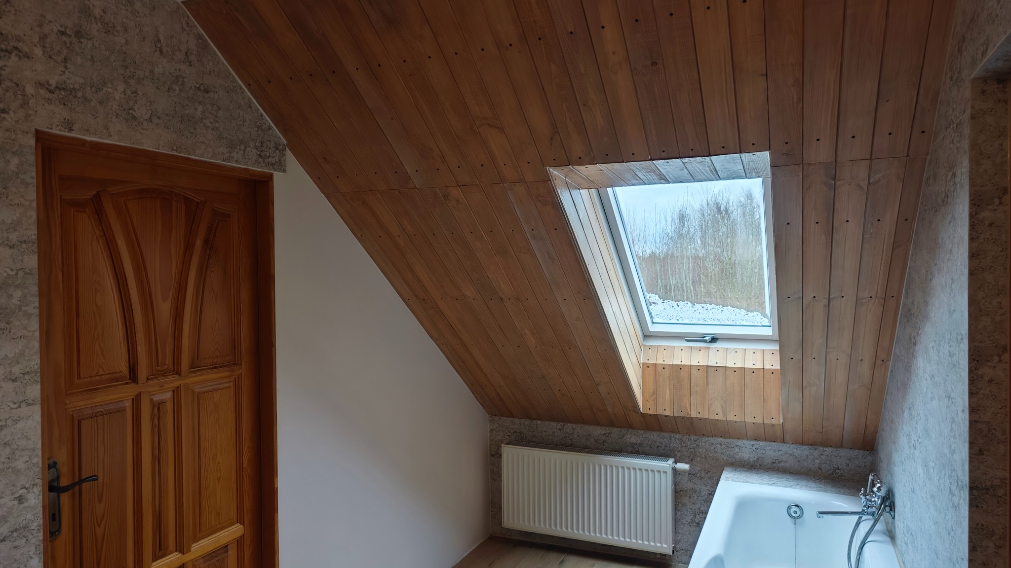 Jasna łazienka na poddaszu ze skośnym drewnianym sufitem, oknem dachowym z widokiem na zimowy krajobraz, drewnianymi drzwiami i grzejnikiem.