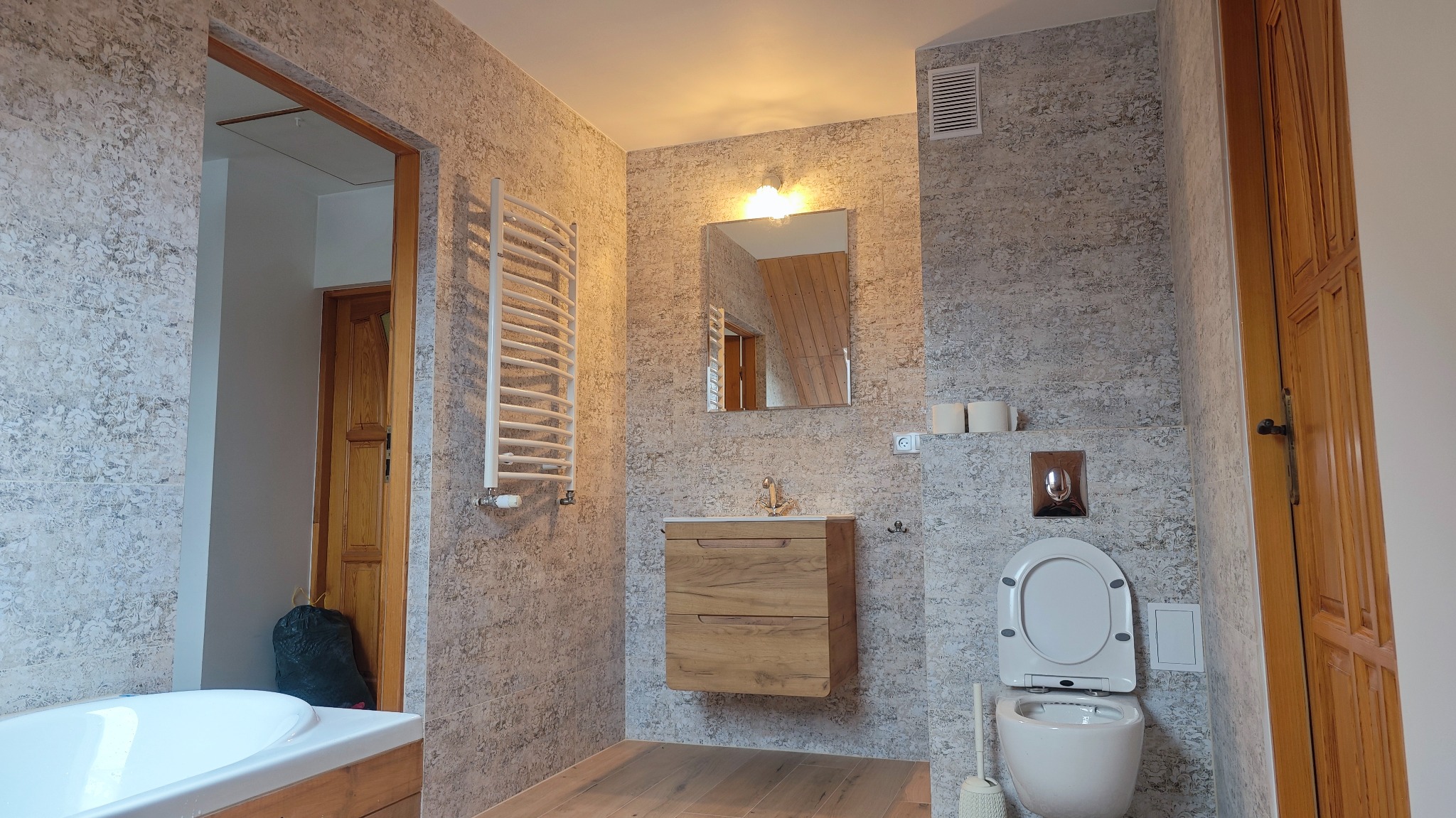 Nowoczesna łazienka z szarymi płytkami imitującymi kamień, drewnianą szafką pod umywalką, grzejnikiem drabinkowym i wiszącą toaletą z chromowanym przyciskiem spłukiwania.