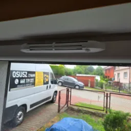 Biały osuszacz kondensacyjny Ventin HY+ zamontowany pod sufitem, widoczny z wnętrza pomieszczenia, przez okno widać samochód dostawczy z reklamą firmy zajmującej się osuszaniem.