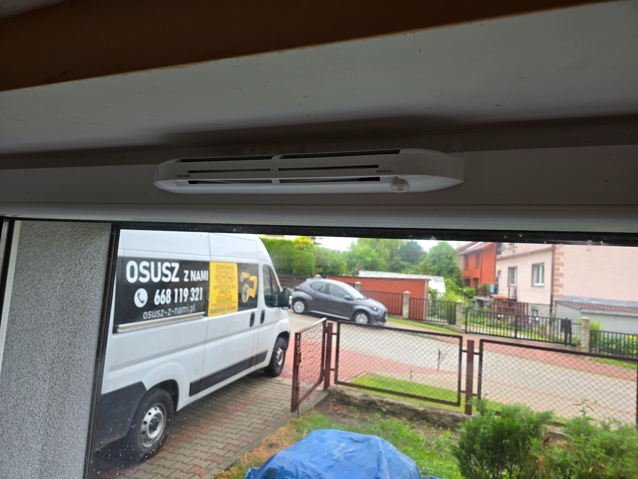 Biały osuszacz kondensacyjny Ventin HY+ zamontowany pod sufitem, widoczny z wnętrza pomieszczenia, przez okno widać samochód dostawczy z reklamą firmy zajmującej się osuszaniem.