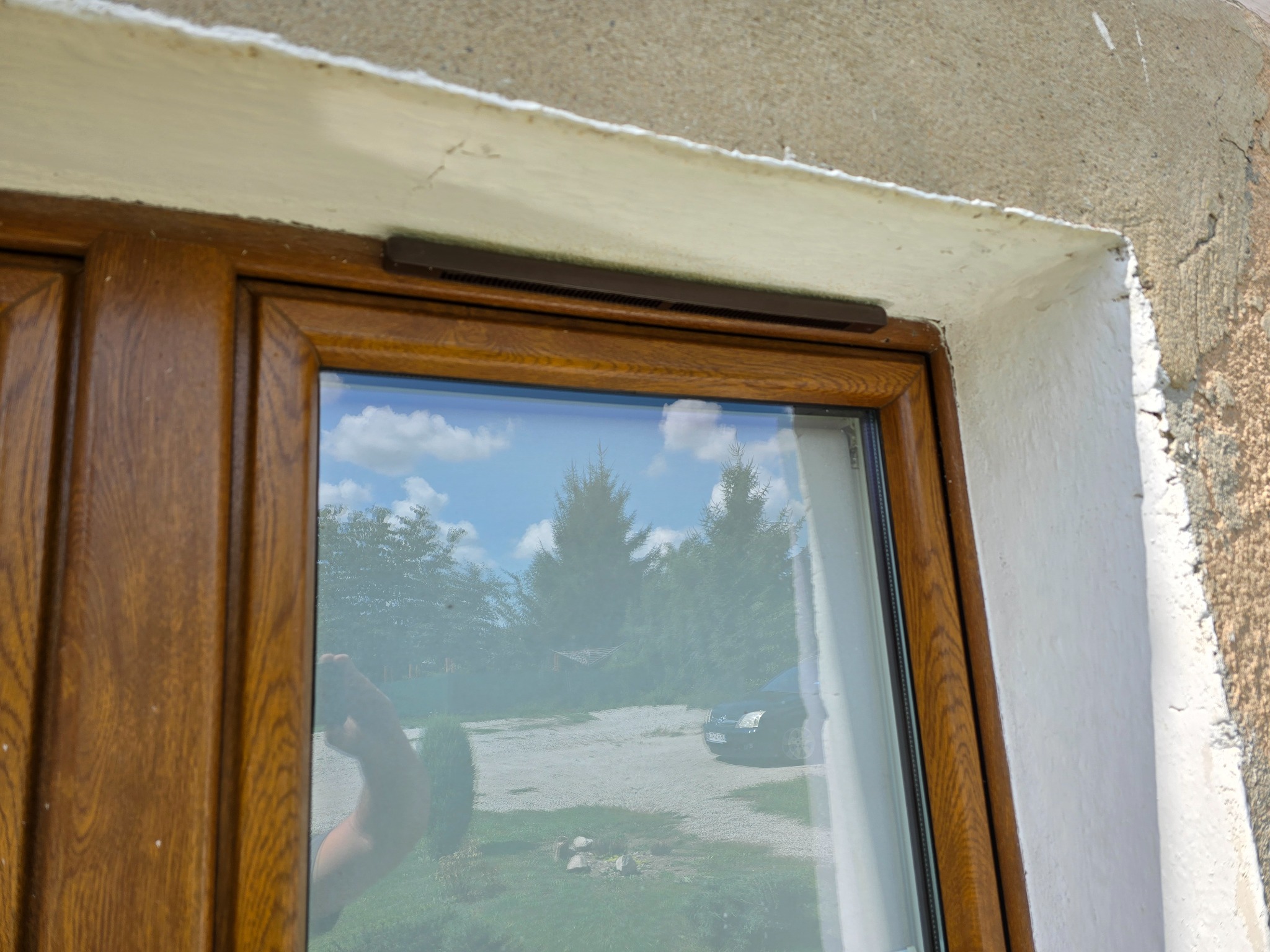 Fragment okna z brązową ramą, widoczny nawiewnik i refleksja otoczenia: drzewa, niebo, samochód.