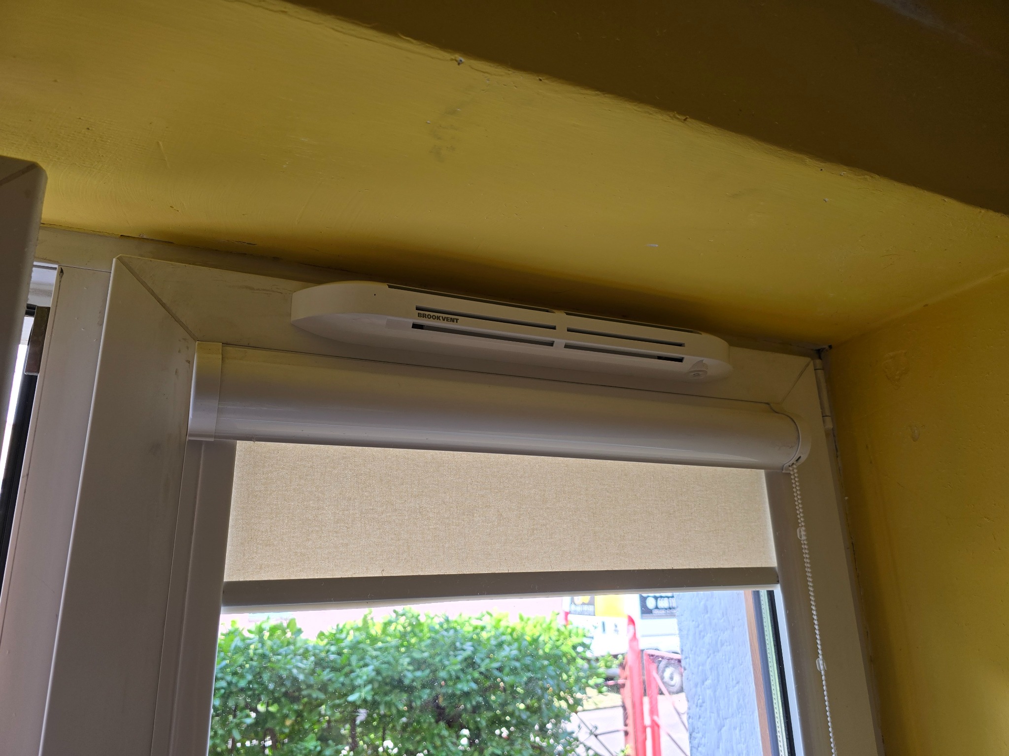 Biały nawiewnik okienny Brookvent zamontowany nad roletą w oknie z widokiem na zieleń na zewnątrz, żółta ściana i sufit w tle.