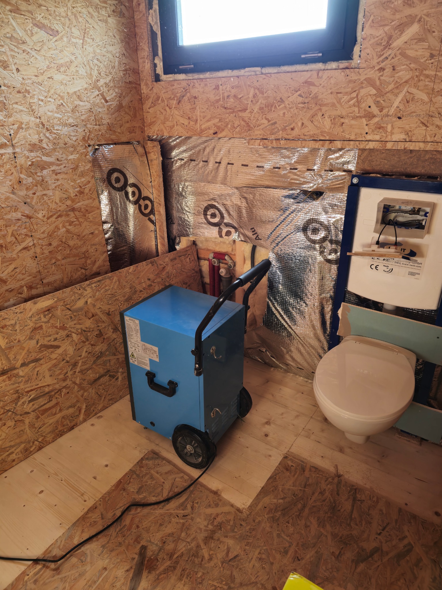 Niebieski osuszacz powietrza na kółkach w surowym wnętrzu łazienki z płyt OSB i izolacją aluminiową, widoczna muszla klozetowa w trakcie montażu.