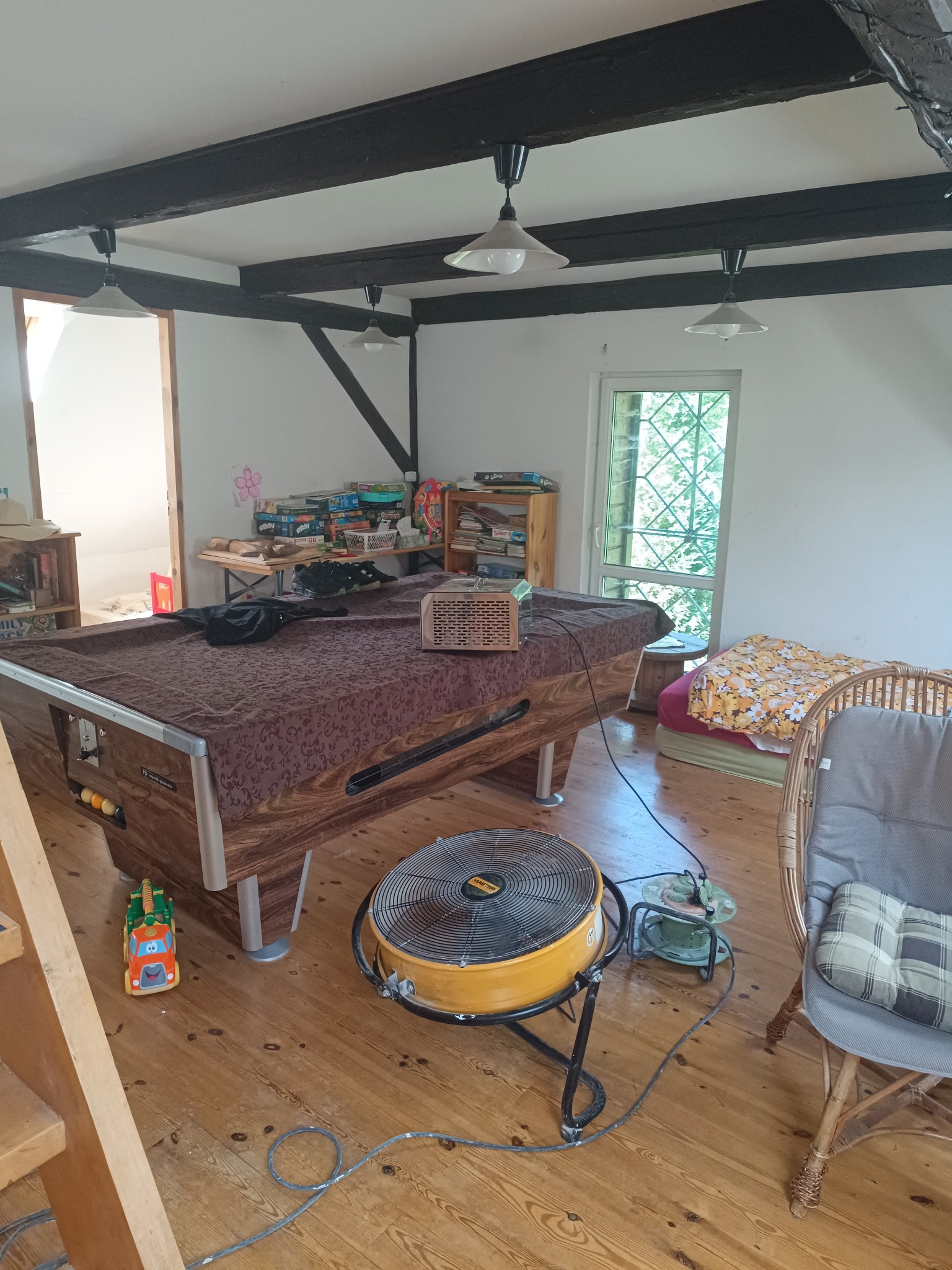 Pokój z drewnianą podłogą i stropem, na środku stół bilardowy, obok żółty wentylator przemysłowy, prawdopodobnie w trakcie procesu suszenia po zalaniu.
