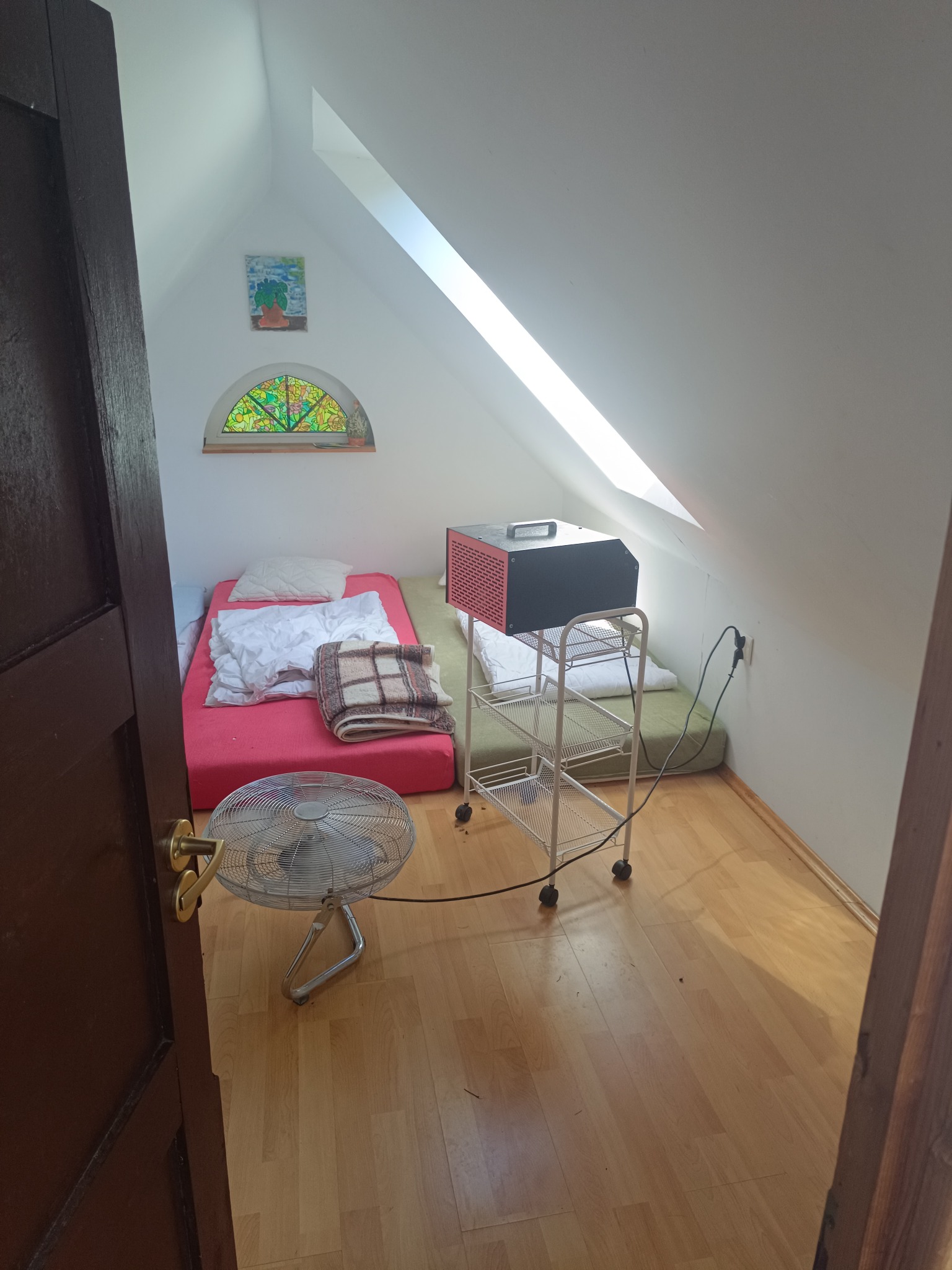 Pokój na poddaszu z dwoma materacami, wentylatorem podłogowym i osuszaczem powietrza na metalowym wózku; widoczne okno ze szprosami i obrazek na ścianie.