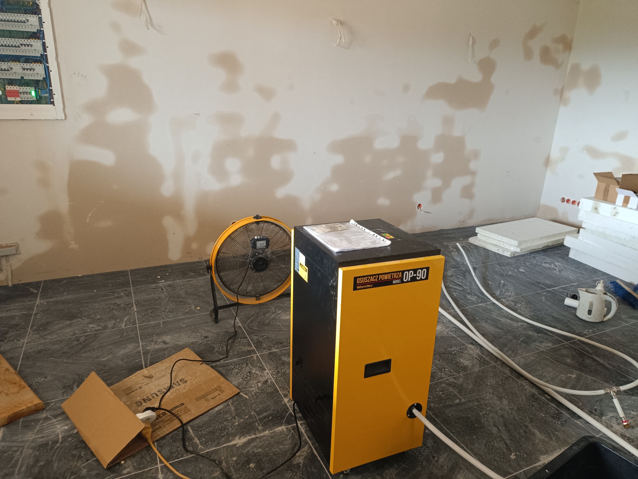Żółto-czarny osuszacz powietrza Warmtec OP-90 w trakcie pracy, widoczne mokre ślady na ścianie, wentylator i elementy instalacji odprowadzającej wodę.