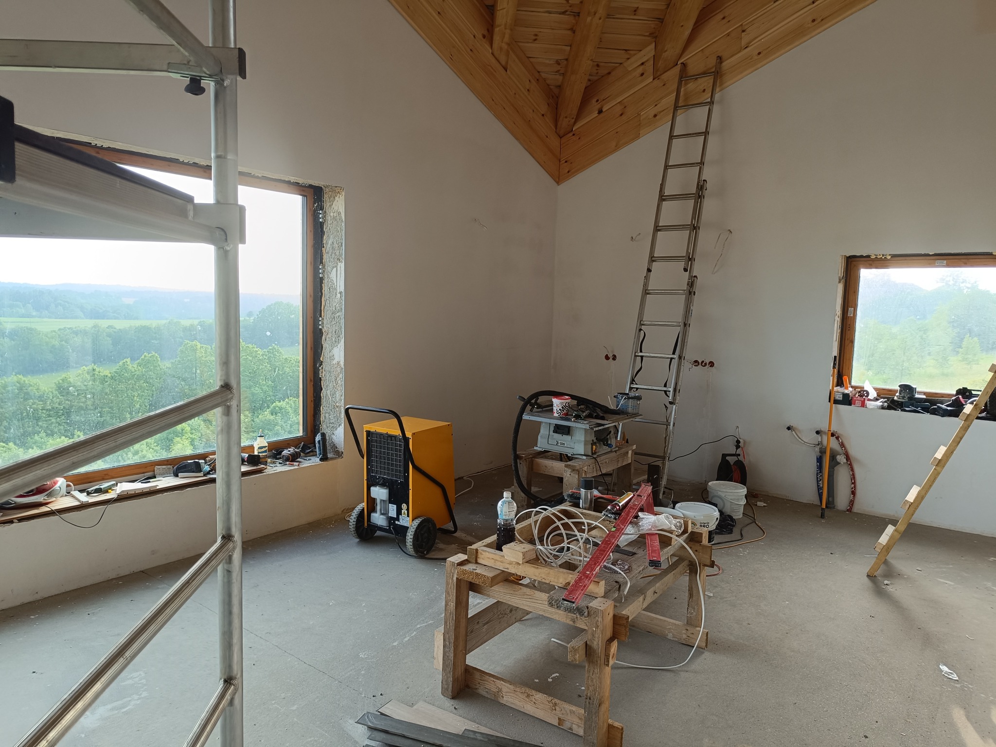 Wnętrze w trakcie remontu z drewnianym stropem, widoczne rusztowanie, drabina oparta o ścianę, żółty osuszacz powietrza na kółkach, stół roboczy z narzędziami i materiałami budowlanymi, okna...