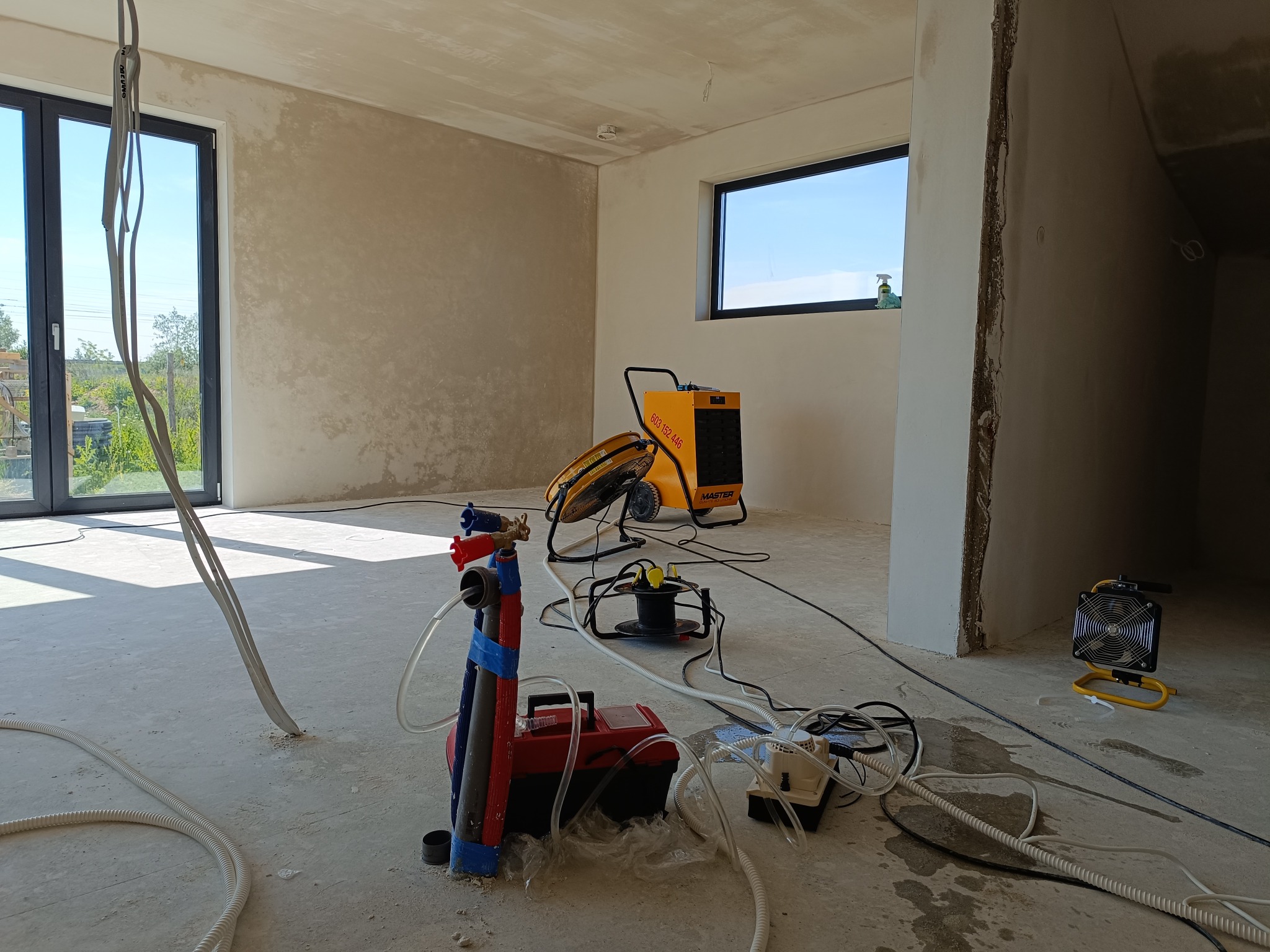 Wnętrze w trakcie prac: żółta nagrzewnica MASTER, pompa z przewodami oraz wentylator na podłodze z widocznymi śladami wilgoci, ściany w stanie surowym.