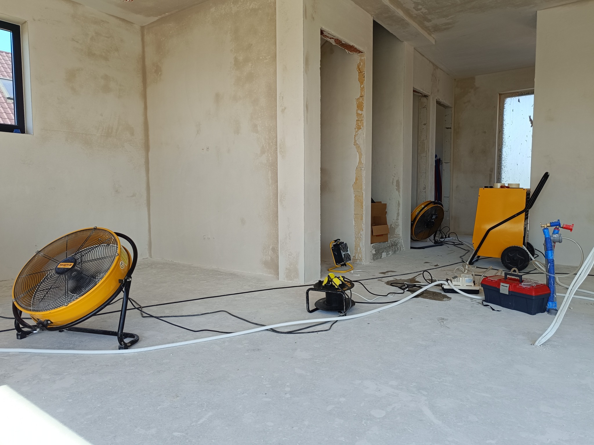 Wnętrze w stanie surowym z widocznymi śladami wilgoci na ścianach, osuszane za pomocą przemysłowych wentylatorów i sprzętu do pomiaru wilgotności, z widoczną skrzynką narzędziową i przewodami...