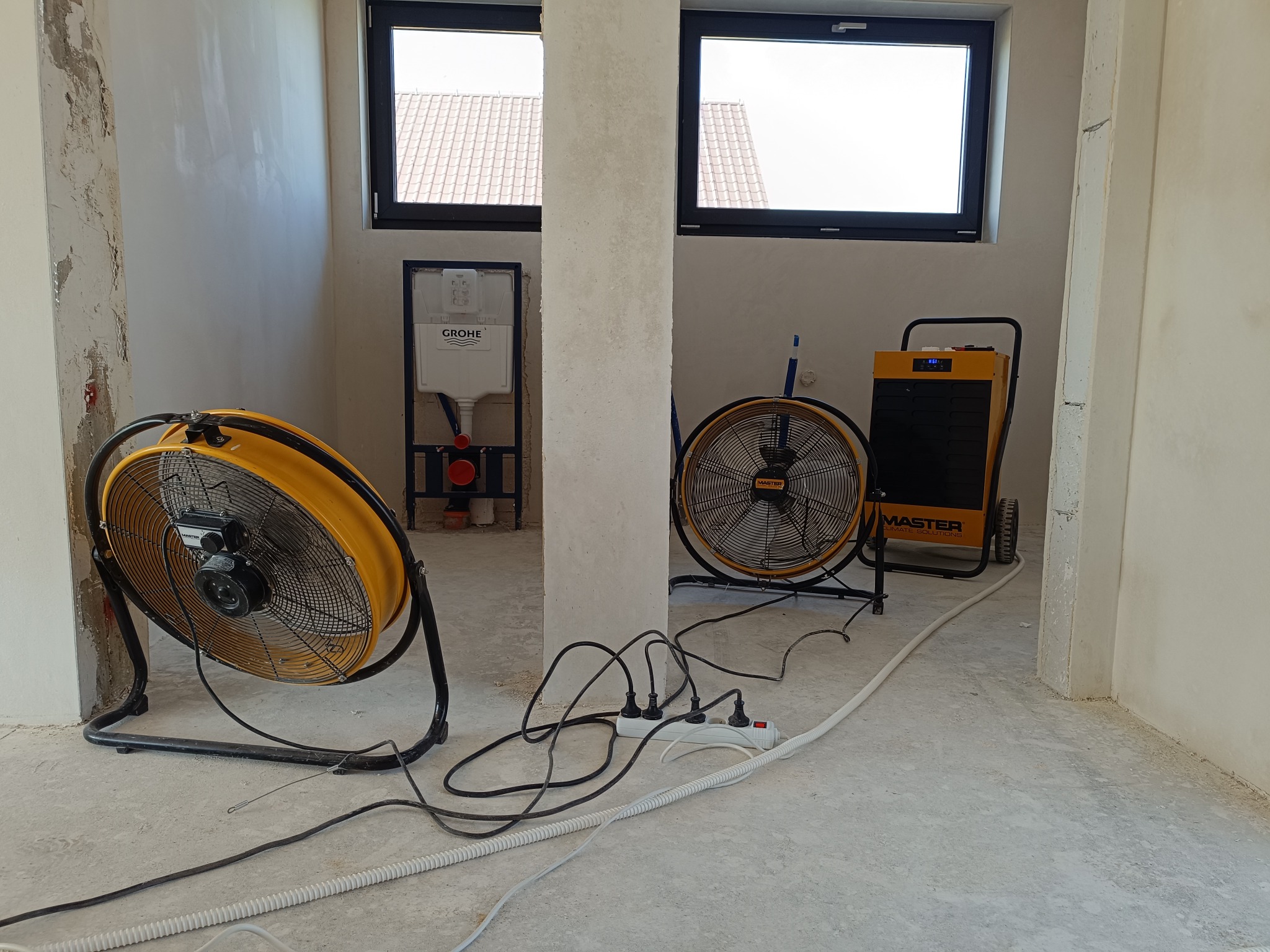 Dwa wentylatory przemysłowe i nagrzewnica Master stojące w surowym wnętrzu z instalacją podtynkową Grohe przed osuszaniem ścian.