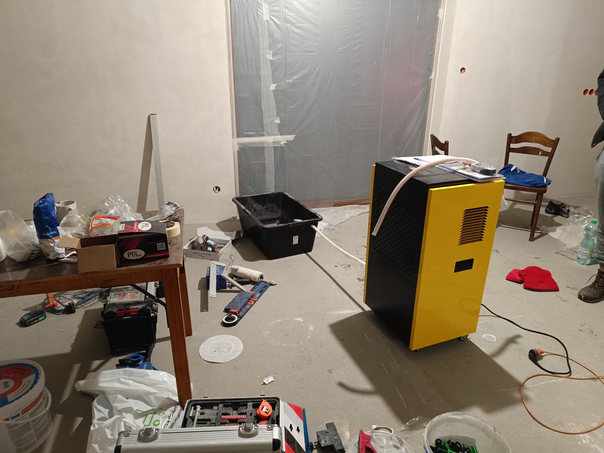 Żółto-czarny osuszacz powietrza z podłączonym wężem stoi w pokoju w trakcie remontu, widoczne zabezpieczone folią okno, narzędzia malarskie, wiadro z farbą i rozłożone narzędzia na stole.