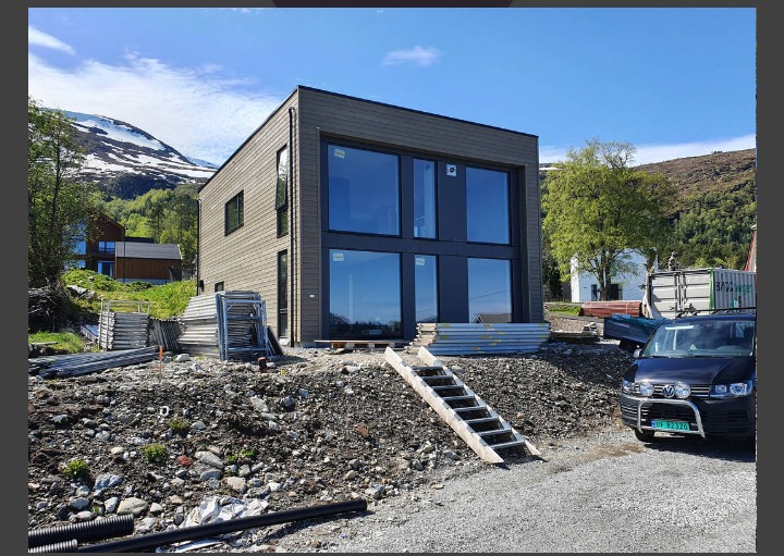 Dwukondygnacyjny, nowoczesny dom o elewacji z drewnianych paneli, z dużymi oknami, w trakcie budowy, z widocznymi rusztowaniami, schodami i samochodem na tle górskiego krajobrazu.