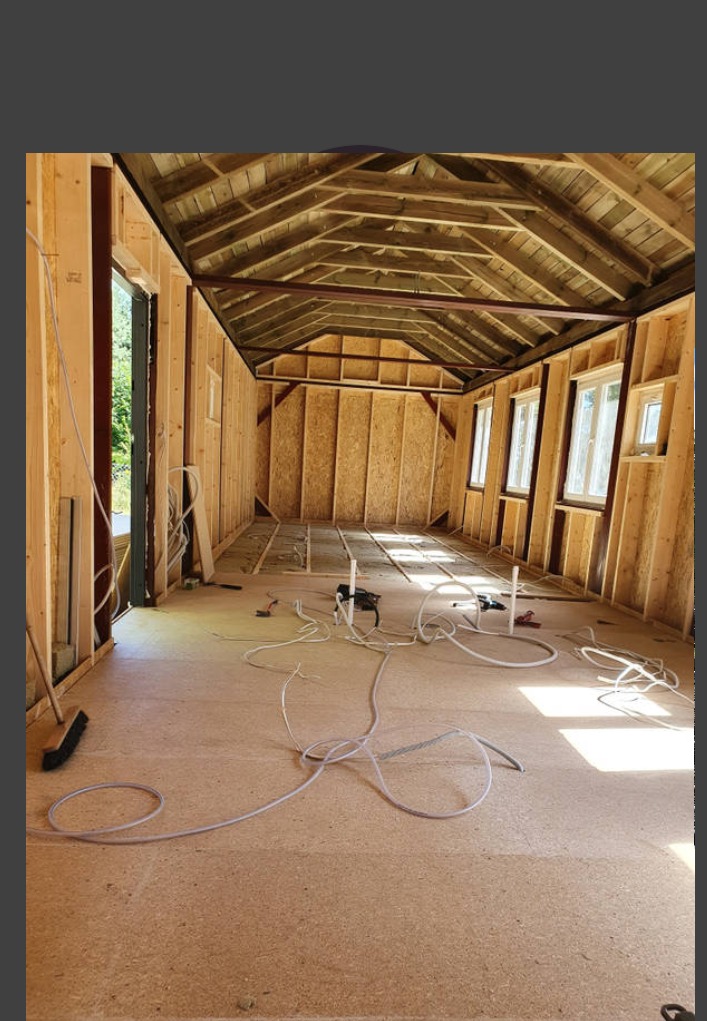 Wnętrze surowego domu w trakcie budowy: drewniana konstrukcja szkieletowa, widoczne okna, drewniany strop, podłoga z płyty OSB, rozłożone kable i narzędzia.