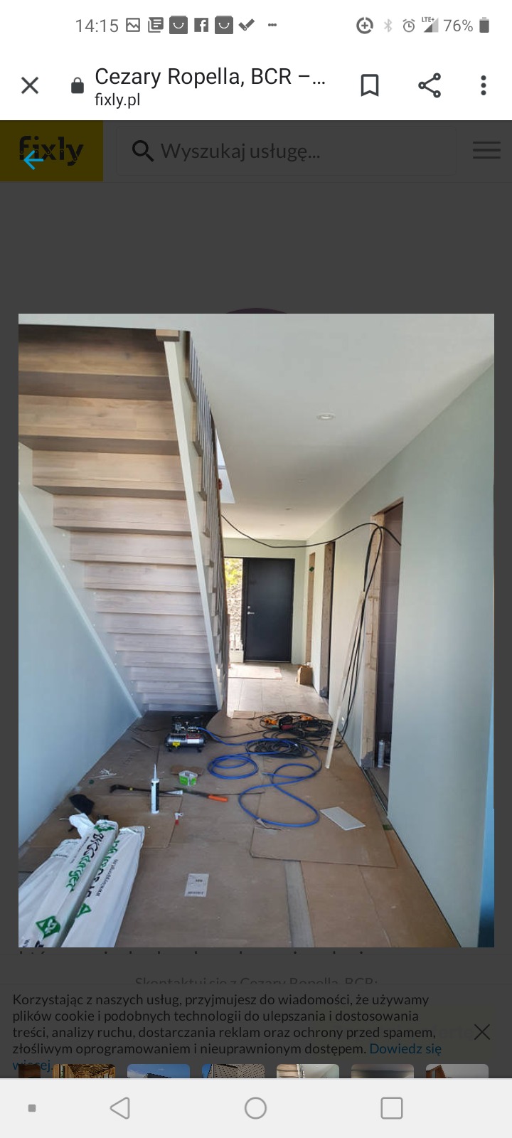 Wnętrze domu w trakcie budowy: widoczne drewniane schody, czarne drzwi wejściowe, narzędzia budowlane i materiały zabezpieczające podłogę.