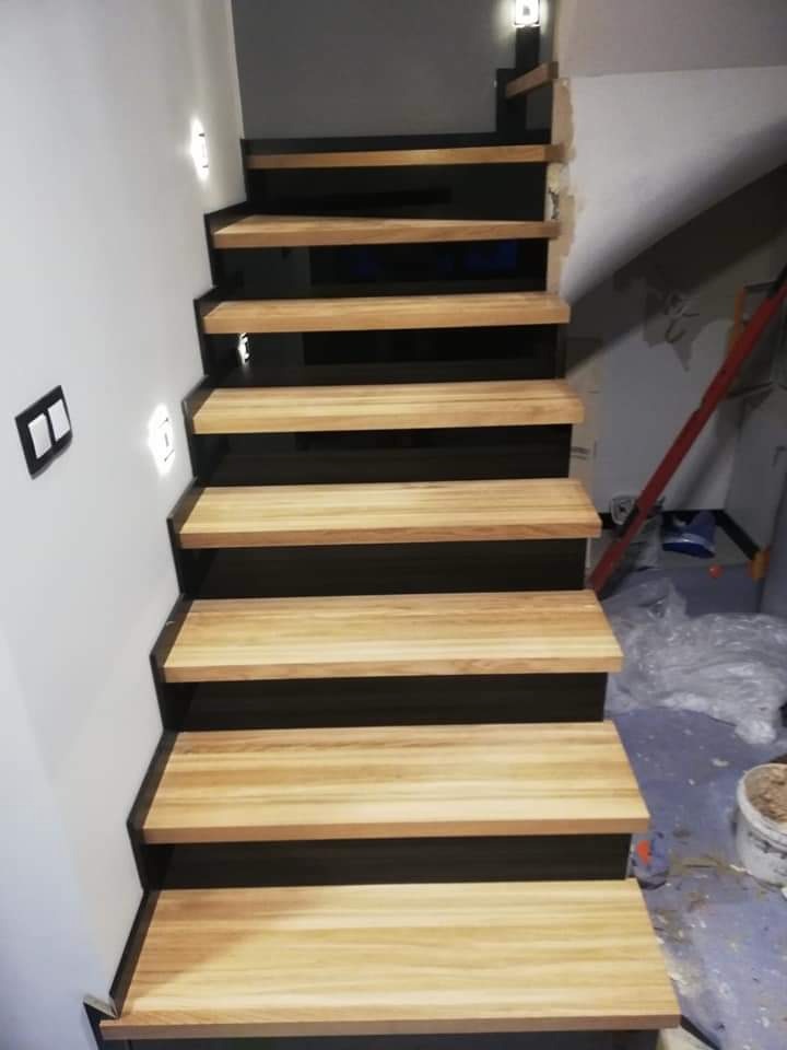 Schody drewniane z czarnymi podstopnicami i oświetleniem punktowym na ścianie, widoczne ślady prac wykończeniowych na ścianie i podłodze.
