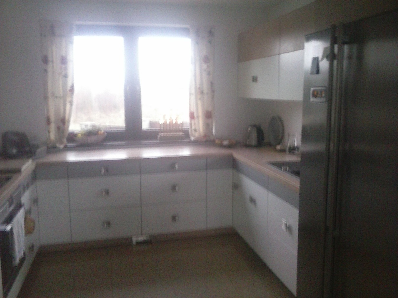 Jasna kuchnia z białymi szafkami z szufladami i stalową lodówką, widok na okno z zasłonami w tle.