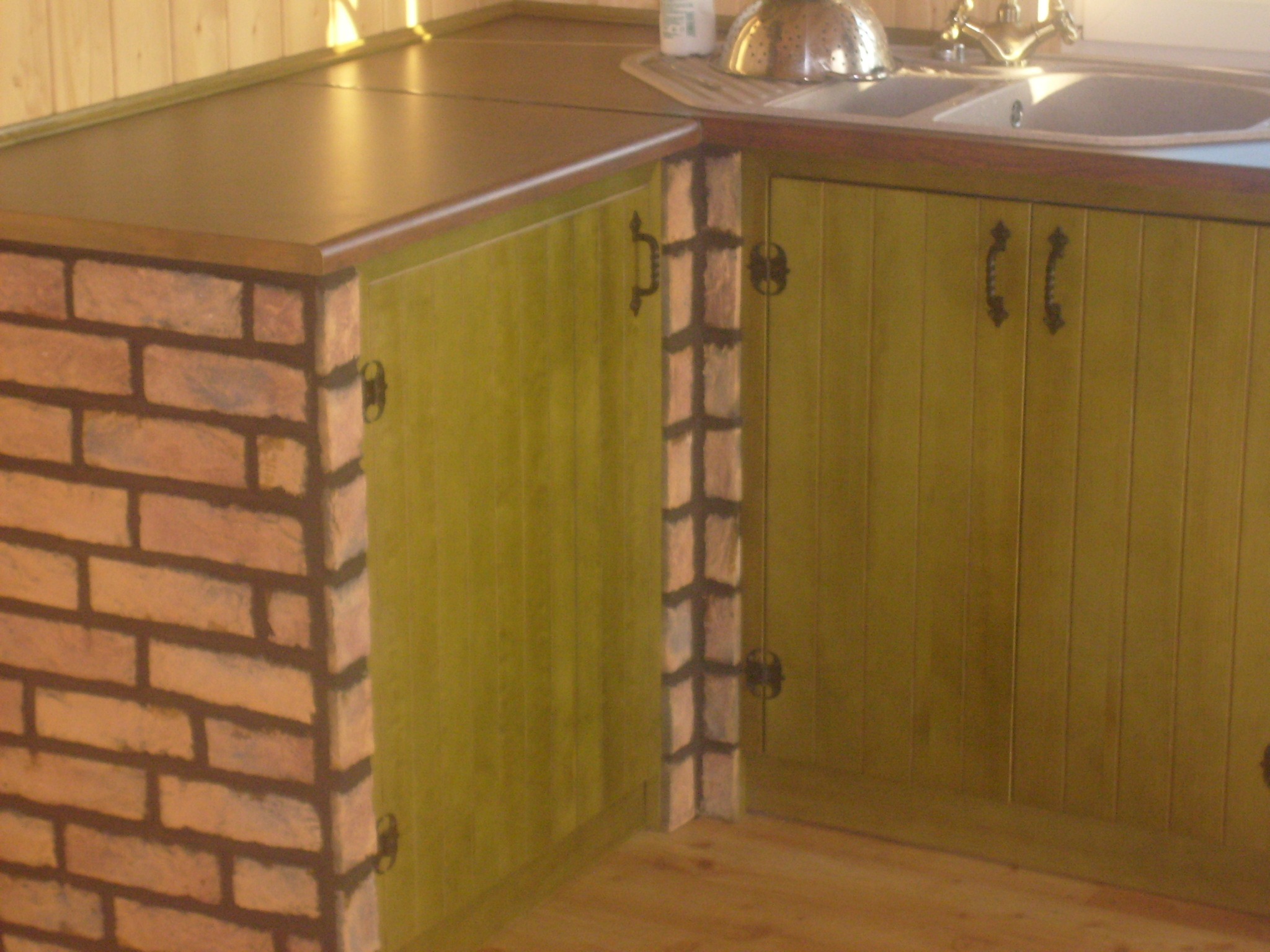 Fragment zabudowy kuchennej z zielonymi szafkami z pionowymi frezowaniami, blatem w odcieniach brązu oraz fragmentem ściany wykończonej cegłą.