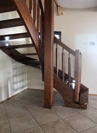 Drewniane schody z ciemnego drewna, z balustradą i słupem nośnym, widoczne na tle jasnych ścian i płytek podłogowych. Schody skręcają w lewo i są oświetlone naturalnym światłem z okna. Widoczny...