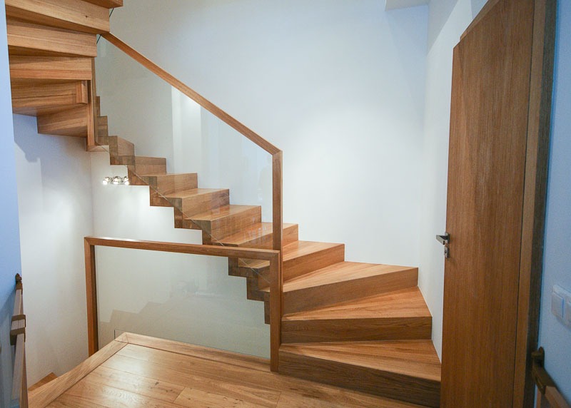 Drewniane schody z przezroczystą balustradą szklaną, oświetlenie punktowe pod schodami, drewniana podłoga i drzwi w tle.