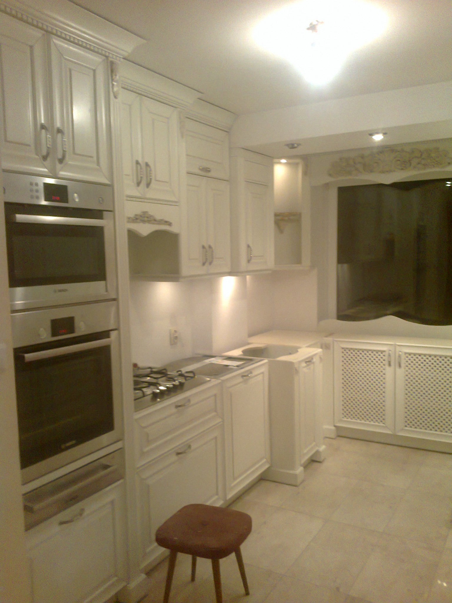 Klasyczna biała kuchnia z frezowanymi frontami, dwoma piekarnikami w zabudowie, gazową płytą grzewczą i małym stołkiem na pierwszym planie.