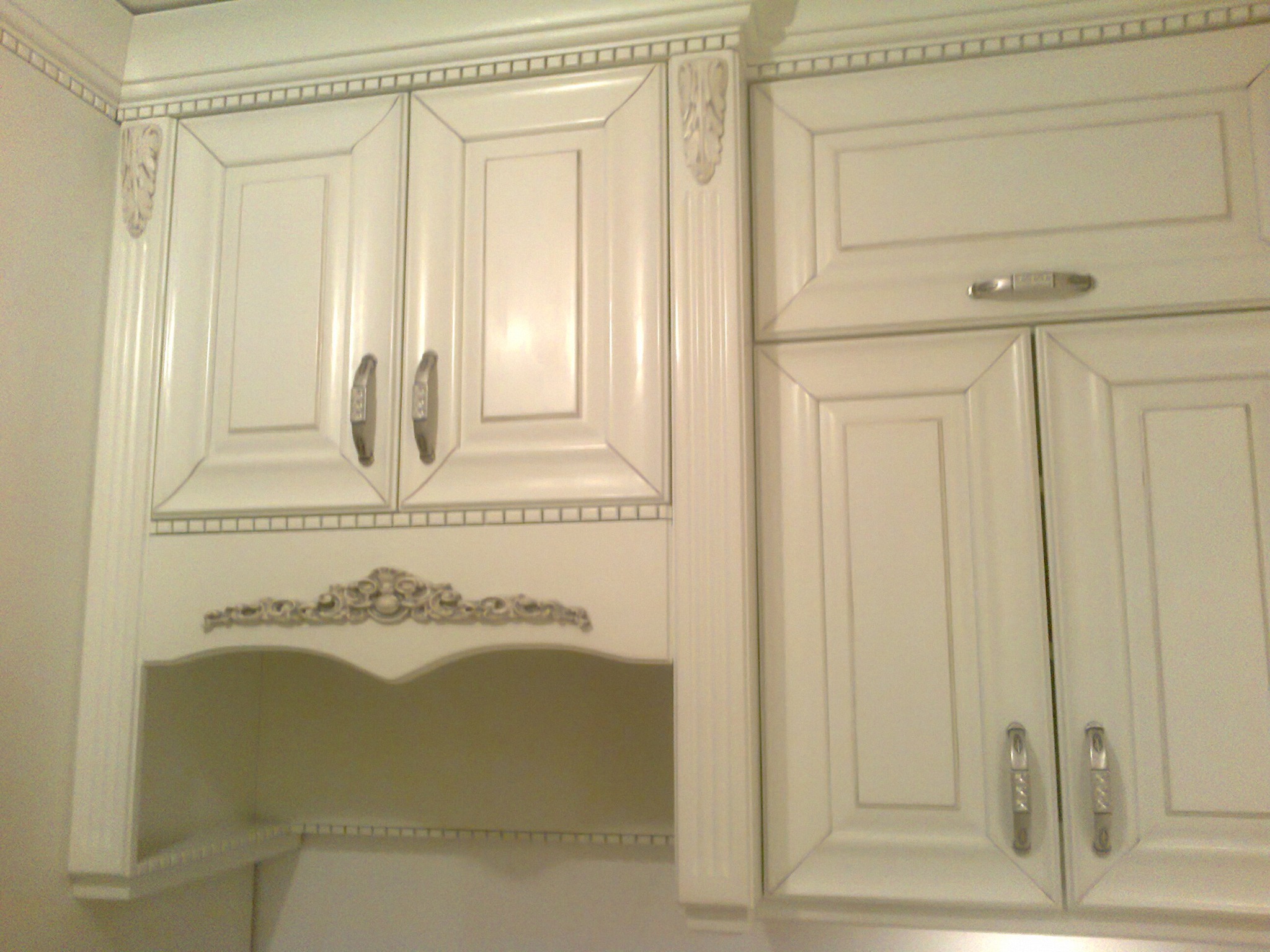 Białe, górne szafki kuchenne z dekoracyjnymi frezowaniami i półką otwartą, oświetlenie delikatnie rozproszone.