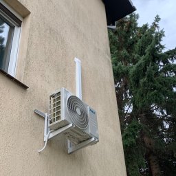 Klimatyczna Sfera Sp. z o.o. - Zewnętrzna jednostka klimatyzacji MDV zamontowana na beżowej elewacji domu, widoczny fragment okna i iglastego drzewa.