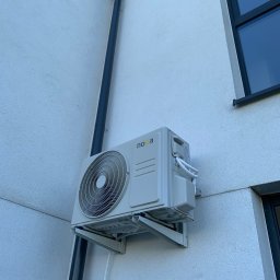 Klimatyczna Sfera Sp. z o.o. - Zewnętrzna jednostka klimatyzacji marki Noxa zamontowana na elewacji budynku mieszkalnego, widoczny czarny spust rynnowy i fragment okna.