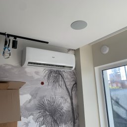 Klimatyczna Sfera Sp. z o.o. - Biały klimatyzator Haier zamontowany na ścianie z tapetą w palmy, widoczne kartony, szyna oświetleniowa i głośnik sufitowy.