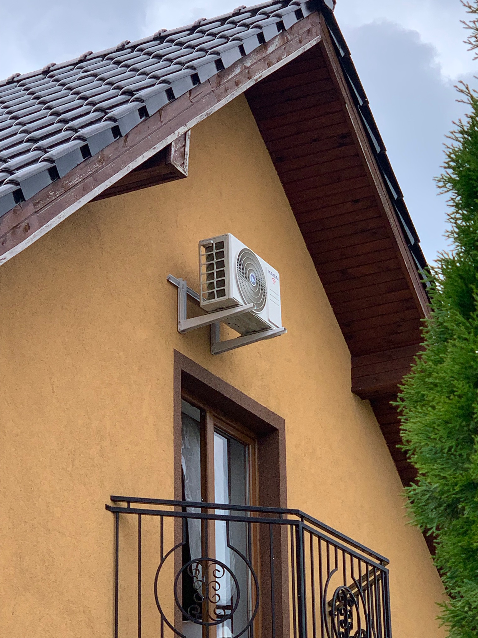 Zewnętrzna jednostka klimatyzacji marki Kaisai zamontowana na ścianie domu z balkonem i brązowym dachem z dachówki ceramicznej.