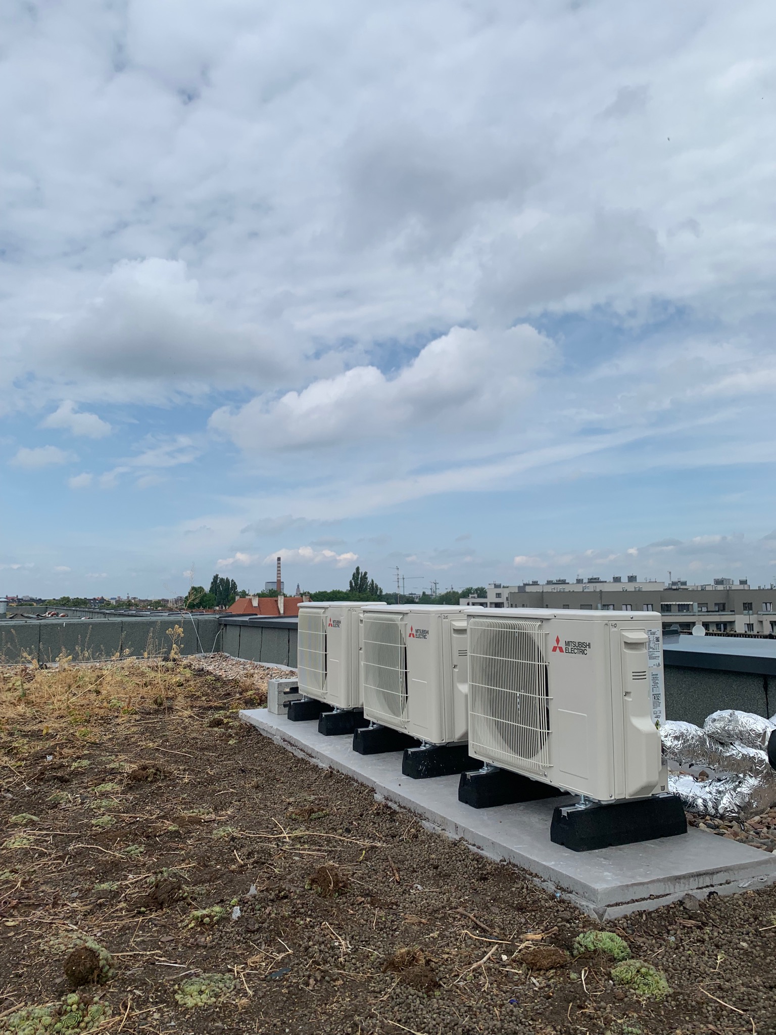Trzy zewnętrzne jednostki klimatyzacyjne Mitsubishi Electric umieszczone na betonowej podstawie na zielonym dachu budynku, widok na miasto w tle pod błękitnym niebem z chmurami.
