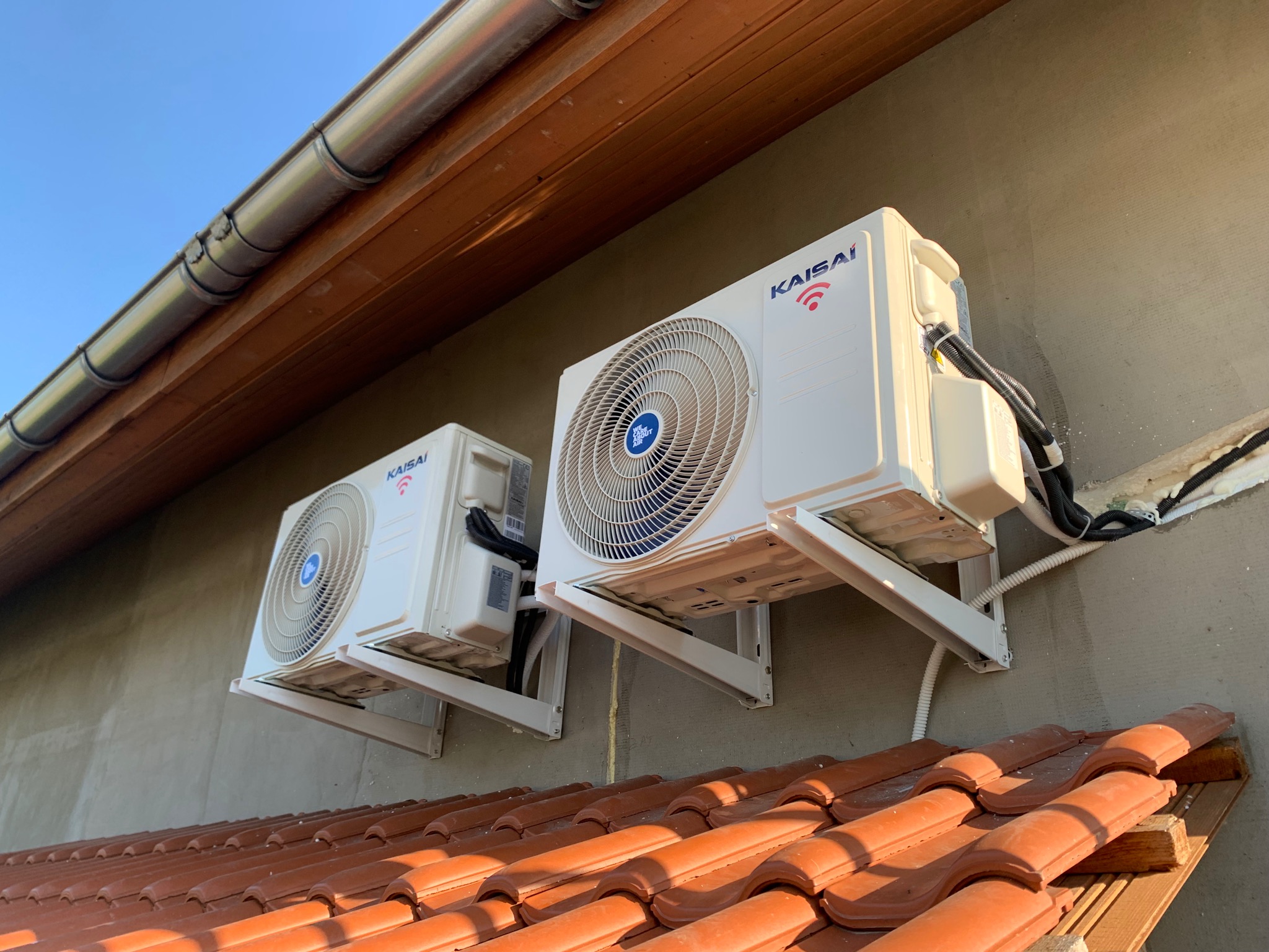 Dwa zewnętrzne moduły klimatyzacji Kaisai zamontowane na ścianie budynku, widoczne kable i rury, dachówka w tle.