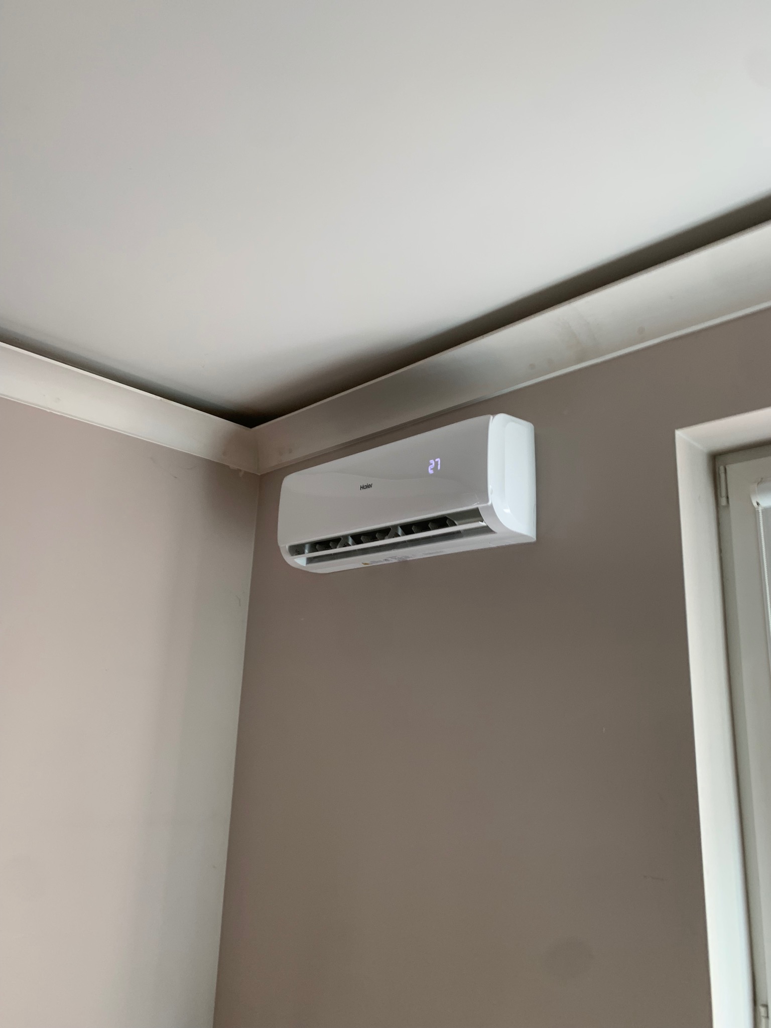 Biały klimatyzator Haier zamontowany na beżowej ścianie w pomieszczeniu z białym sufitem i listwą sufitową.