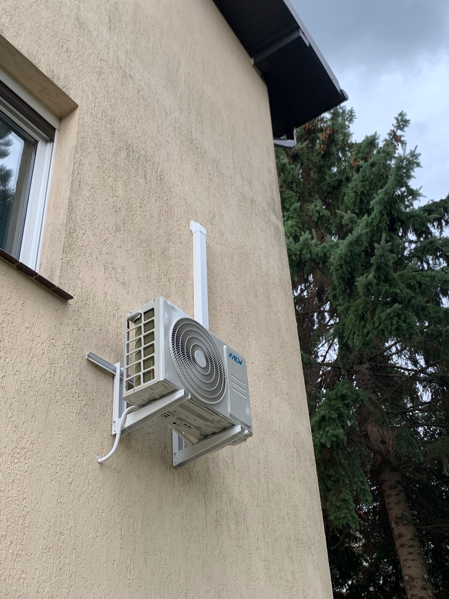 Zewnętrzna jednostka klimatyzacji MDV zamontowana na beżowej elewacji domu, widoczny fragment okna i iglastego drzewa.