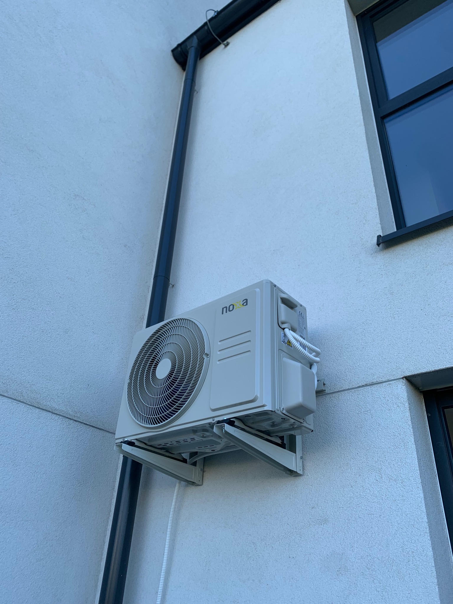 Zewnętrzna jednostka klimatyzacji marki Noxa zamontowana na elewacji budynku mieszkalnego, widoczny czarny spust rynnowy i fragment okna.