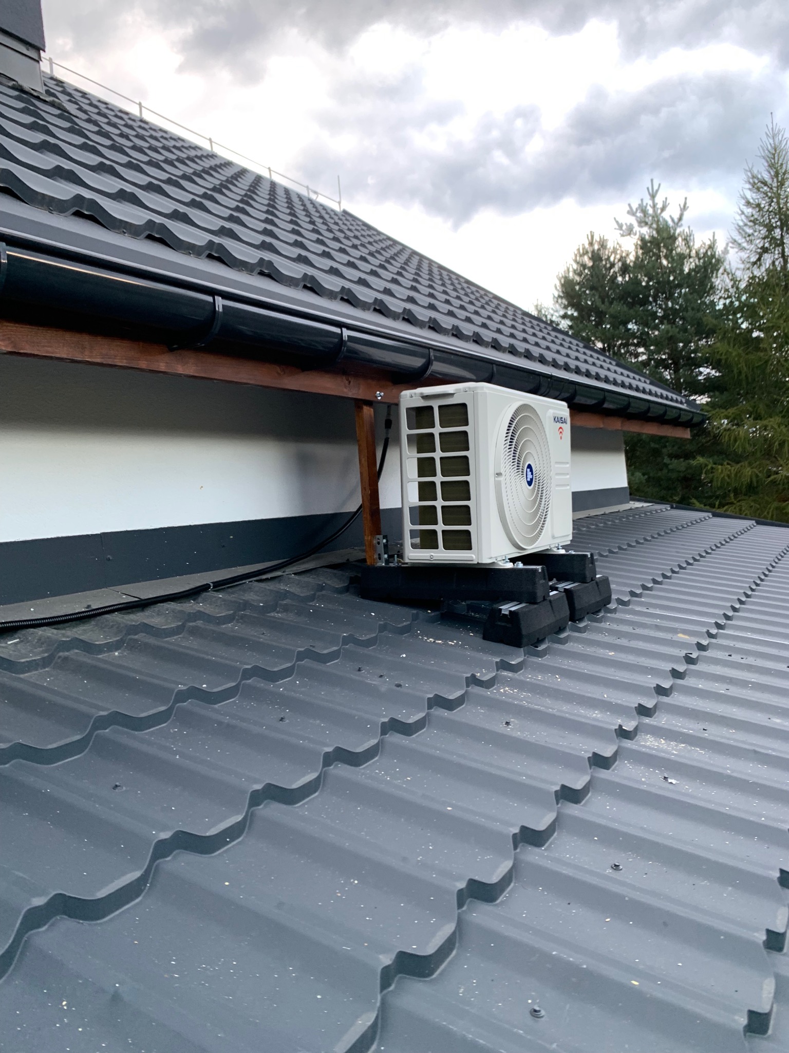 Biała jednostka zewnętrzna klimatyzacji marki KAISAI zamontowana na specjalnej konstrukcji na szarym, metalowym dachu domu jednorodzinnego, widoczne drzewa iglaste w tle i pochmurne niebo.