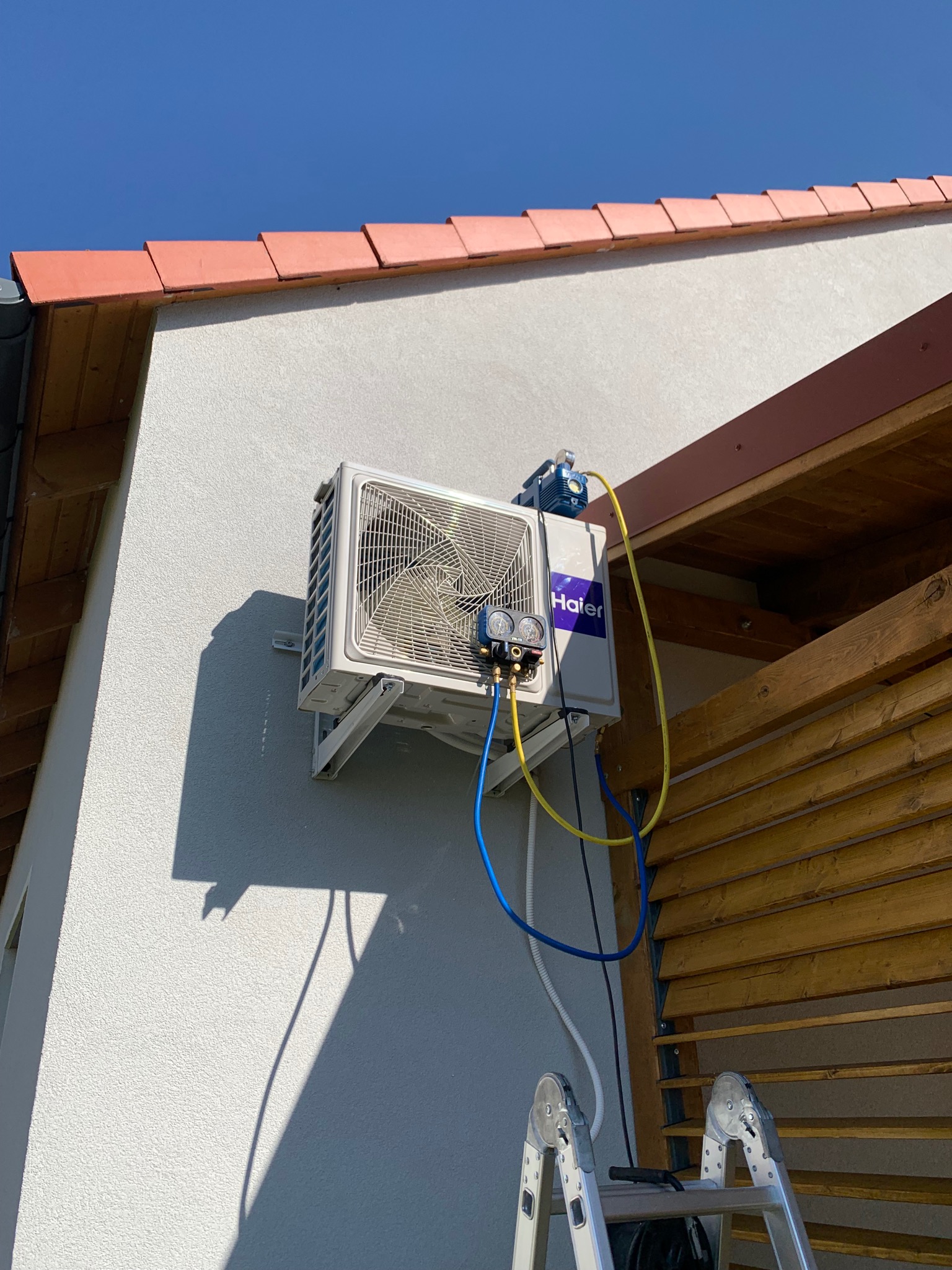 Zewnętrzna jednostka klimatyzacji Haier zamontowana na ścianie domu, podłączona do pompy próżniowej i manometrów serwisowych, widoczna drabina.
