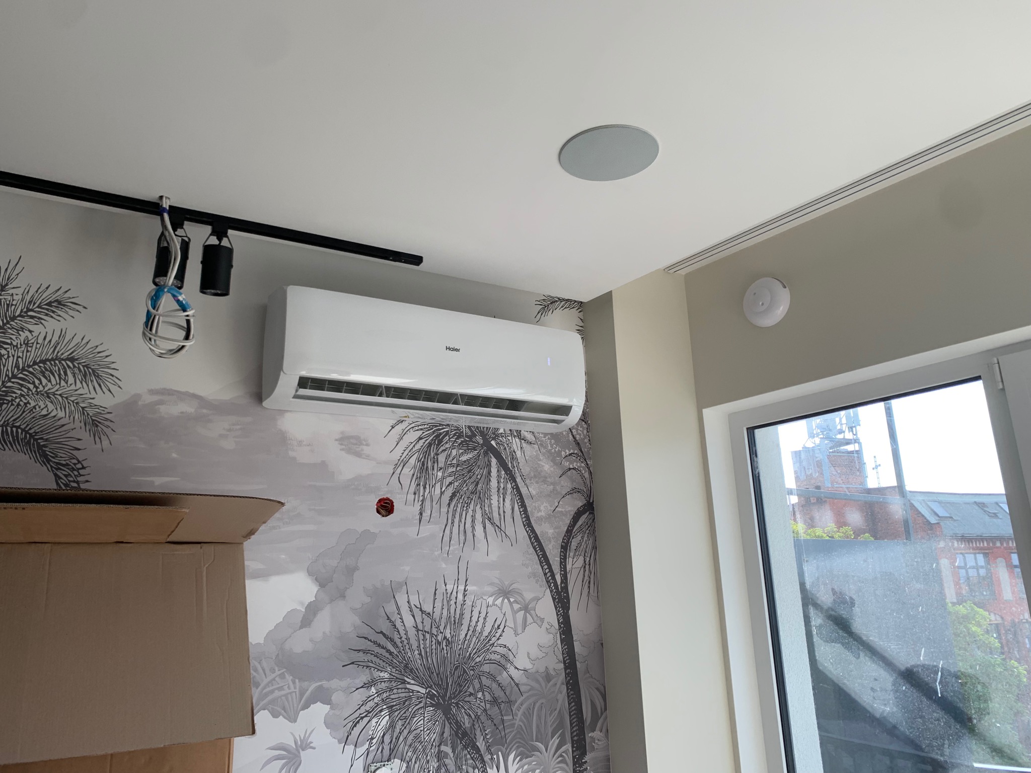 Biały klimatyzator Haier zamontowany na ścianie z tapetą w palmy, widoczne kartony, szyna oświetleniowa i głośnik sufitowy.