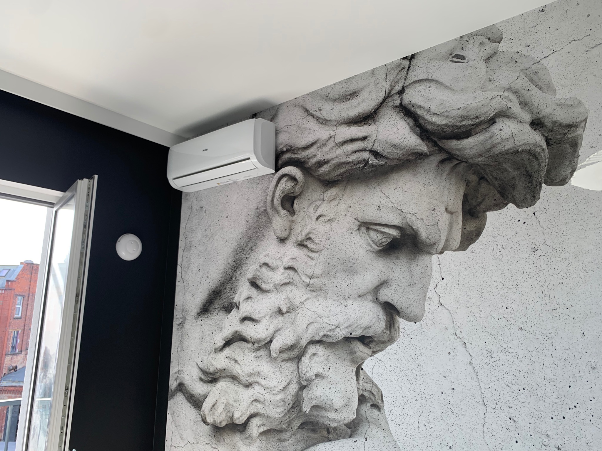 Biały klimatyzator Haier zamontowany na ścianie z fototapetą przedstawiającą popiersie rzeźby, widok z dołu.