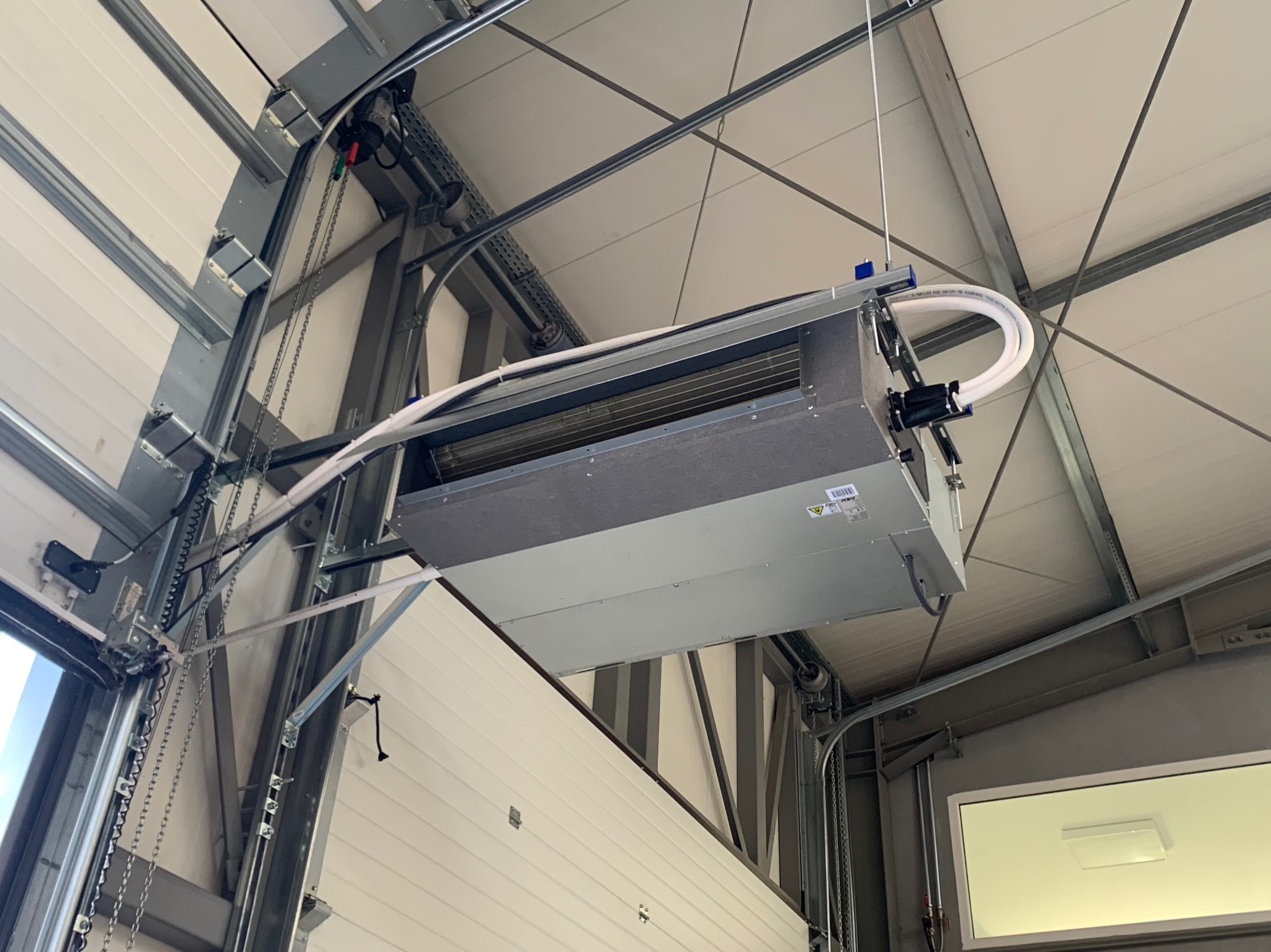 Klimatyzator kasetonowy zamontowany pod sufitem w pomieszczeniu przemysłowym z widocznymi elementami konstrukcyjnymi sufitu i bramą garażową.