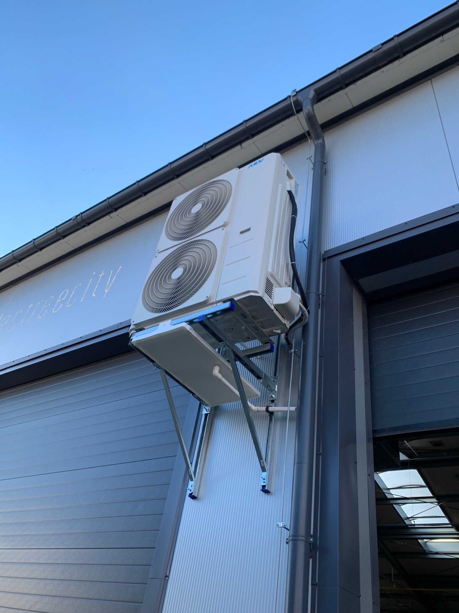 Zewnętrzna jednostka klimatyzacji Daikin zamontowana na metalowej konstrukcji na ścianie budynku przemysłowego z widocznym napisem 'electricity'.
