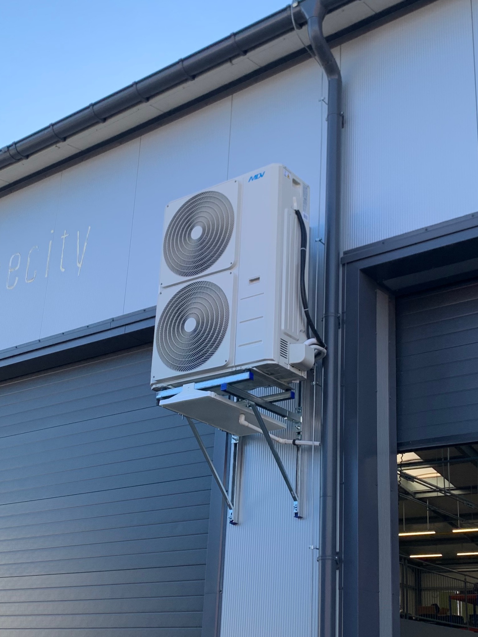 Biała jednostka zewnętrzna klimatyzacji z dwoma wentylatorami, zamontowana na metalowym wsporniku na elewacji budynku przemysłowego z widoczną rynną spustową.