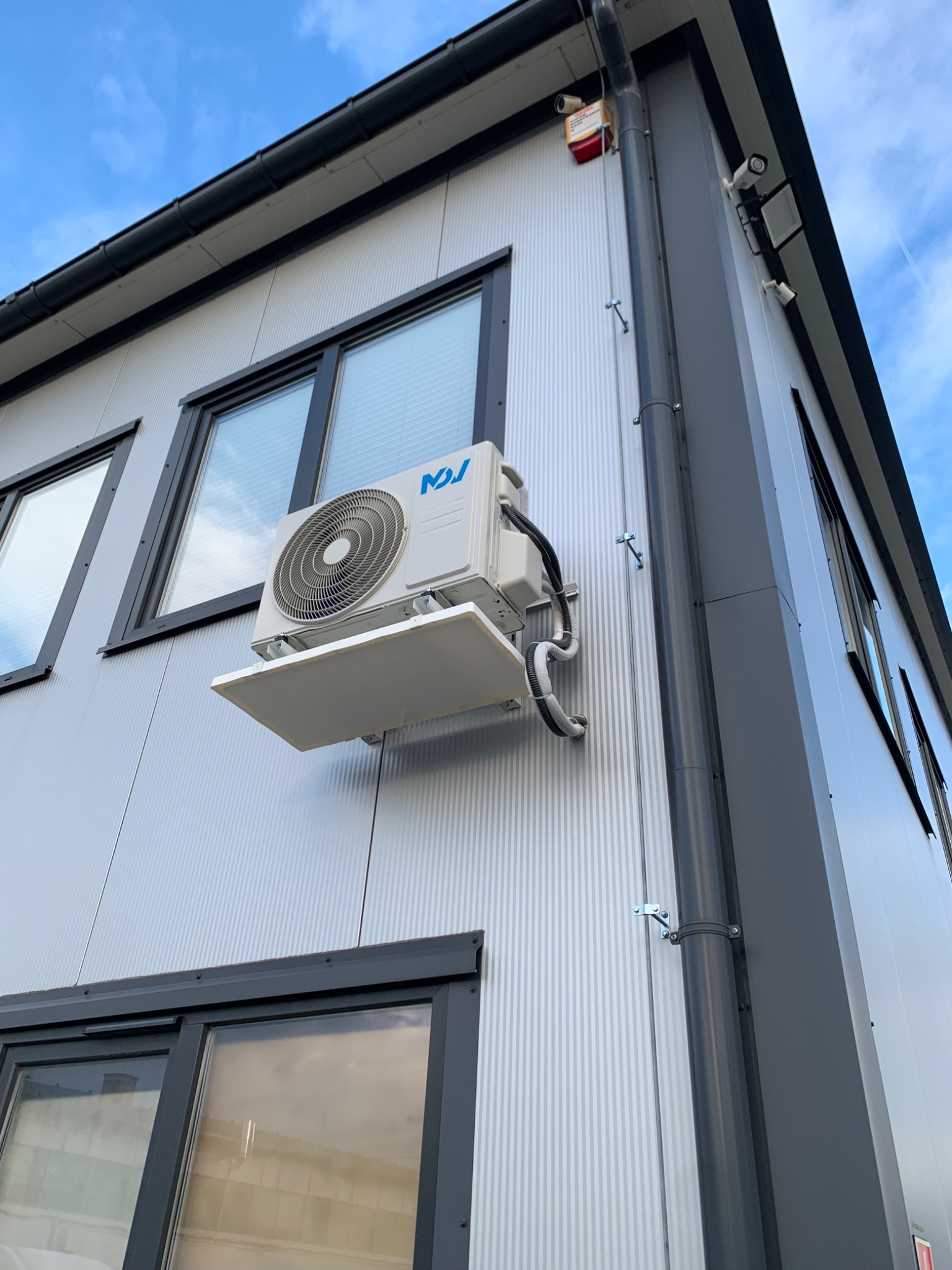 Zewnętrzna jednostka klimatyzacji biurowej zamontowana na metalowej ścianie budynku z widocznymi oknami i systemem rynnowym.