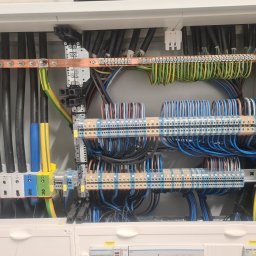 PROINSTAL - Rozdzielnia elektryczna z widocznymi przewodami o różnych kolorach, listwami zaciskowymi i elementami montażowymi, z naklejkami informacyjnymi w dolnej części.